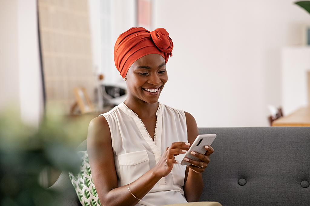 Woman in head scarf smiling as she sits on sofa and looks at her phone