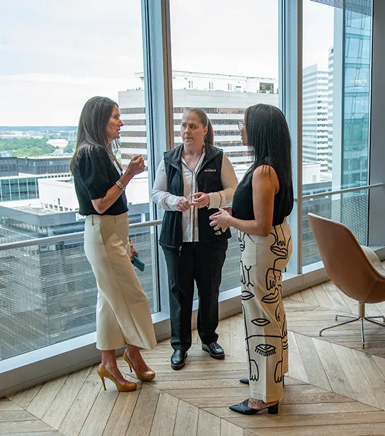 Three female employees talking collaboratively in the Evolent office near large windows, with office buildings in the background